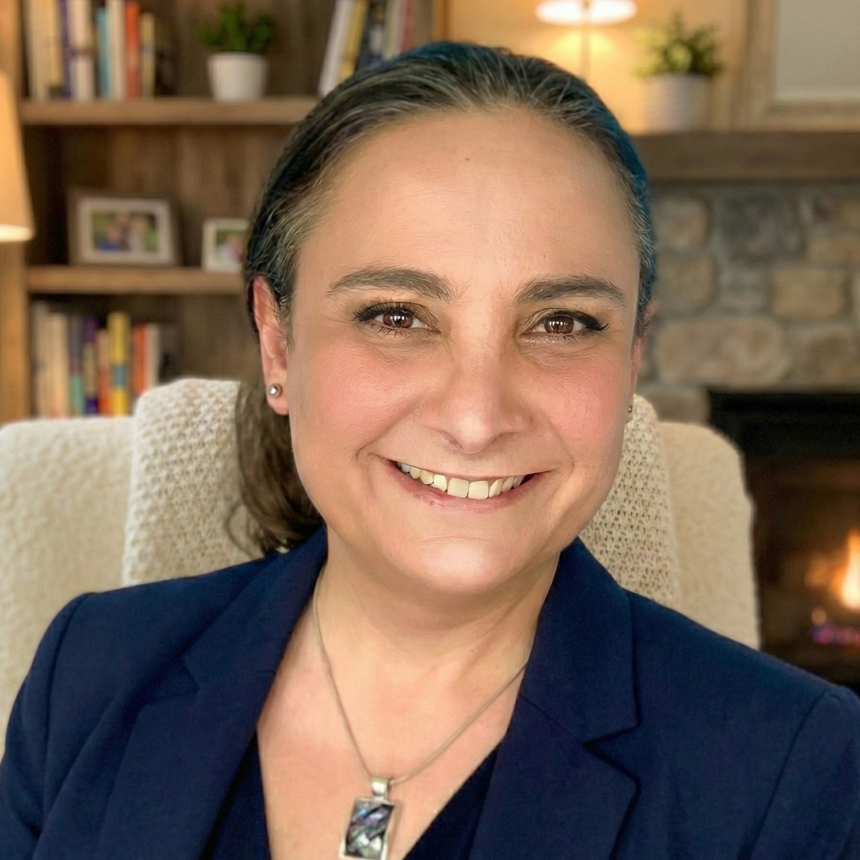 A professional portrait of the founder, a smiling woman with dark hair tied back, wearing a blue blazer and an abalone shell necklace, seated in front of a warm fireplace and bookshelf.